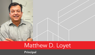 A man with short hair smiles confidently while leaning against a wall, with a friendly demeanor and professional attire. Red and gray branding is visible.