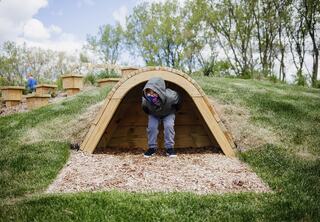 A child peeks out from a wooden playground feature in a grassy, hilly area, surrounded by trees and playful structures, enjoying the outdoors at Prairie Trails School's natural playground.