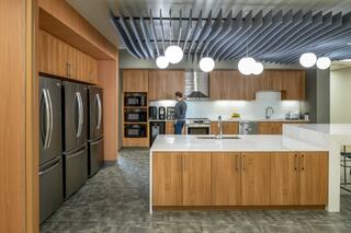 The second floor of Downers Grove Civic Center features a shared kitchen with sleek wooden cabinetry, stainless steel appliances, and a central island, with a person engaged in meal prep.