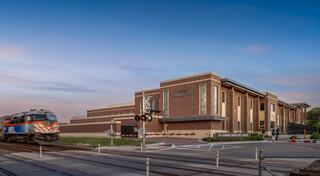 The new Downers Grove Civic Center stands beside the Metra railroad tracks at sunset, with a train approaching and a signal light illuminated nearby.