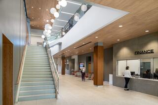 A Bright two-story atrium and lobby at the Downers Grove Civic Center features a staircase, wooden accents, and a finance desk, with soft lighting and seating areas for visitors.