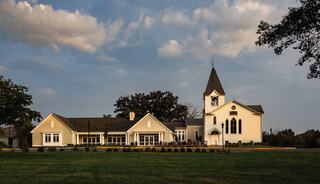 A charming white church with a tall spire stands beside a modern building, surrounded by lush greenery and a cloudy sky.