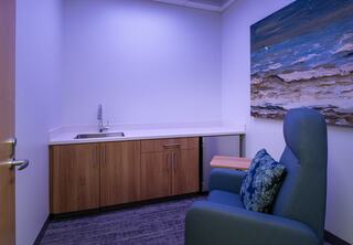 A small quiet room or mothers room provides peace, calm and quiet for staf in the Downers Grove Civic Center. Room features a cozy arm chair, nature-inspired wall art, countertop and cabinets with a sink and mini fridgel