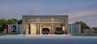 Front facade of the new Channahon Fire Protection District Station No. 2 photographed at dusk. Three apparatus bays are illuminated from inside. Personnel are shown inside the bays next to an ambulance and fire engine.