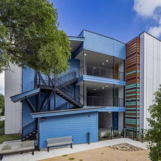 A modern affordable housing features a vibrant, colorful side, with a stairway leading to balconies amidst green trees and a landscaped area.