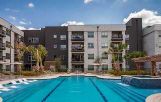 Modern apartment complex featuring a vibrant pool area, lounge chairs, and palm trees, set against a clear blue sky.