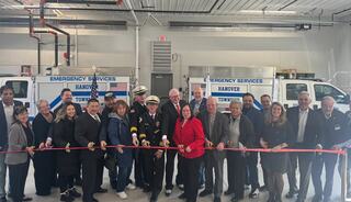 A diverse group gathers to cut a ceremonial ribbon in front of emergency service vehicles, celebrating a new facility.