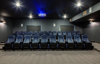 A darkened cinema room features rows of comfortable blue seats facing a projector and a blank screen, surrounded by soft lighting.