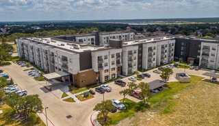 A modern apartment complex with multiple stories, surrounded by green grass, parking spaces, and trees, under a partly cloudy sky.