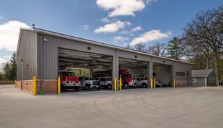 A modern fire station with multiple garage doors, housing several emergency vehicles. Surrounding area features trees and a clear blue sky.