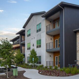 Independent living three-story apartment building with a mix of materials, green landscaping, and balconies, set against a clear blue sky.