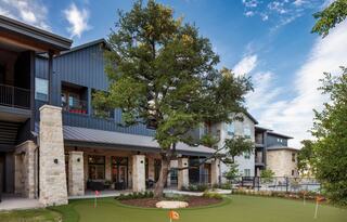Independent living apartment complex with stone accents features a lush green lawn, a large tree, and a bright blue sky in the background.