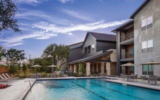 A modern pool area with lounge chairs and umbrellas, surrounded by lush greenery and stylish apartments under a scenic sky.