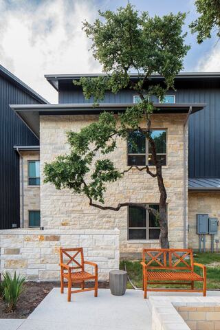 A modern outdoor space features two wooden chairs beside a stone wall, with a unique tree growing alongside a contemporary building.