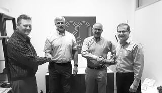 Four men are shaking hands, smiling, and standing together in an office setting, with a logo in the background celebrating 20 years.