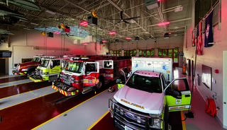 A modern fire station features multiple emergency vehicles, including fire trucks and an ambulance, with red lighting enhancing the interior.