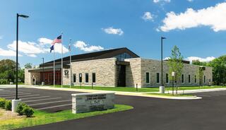 A modern fire station features a stone facade, large windows, and a well-maintained landscape, with flags flying in front under a blue sky.