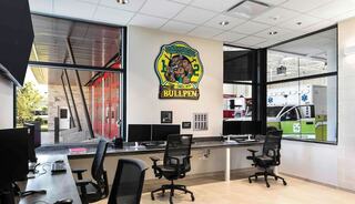 A modern fire station office and watch room features sleek desks and chairs, large windows with a view of emergency vehicles, and a vibrant wall mural.