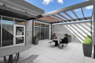 A modern outdoor patio features a table with two people seated, surrounded by planters and a pergola, under a bright blue sky.