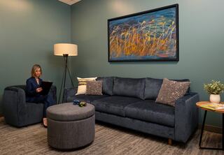 A stylish, calming room features a dark blue couch, a round ottoman, and an artistic painting, with a woman sitting and reading nearby.