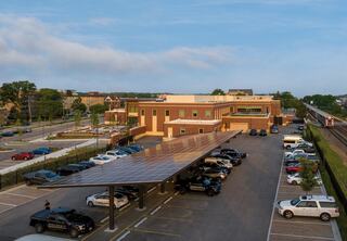 A modern building with a solar panel canopy covers parked police vehicles, surrounded by green space and the nearby Chicagoland Metra train track. Clear blue skies above.