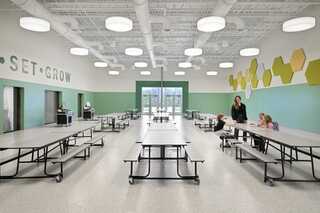 A bright and spacious cafeteria features modern furniture, hexagonal wall decor, and children interacting with an adult in a nurturing environment.