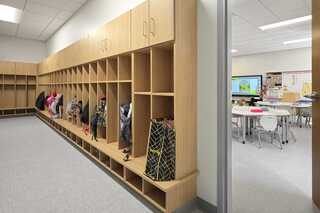 Wooden cubbies line a hallway, filled with colorful backpacks, leading into a bright, organized classroom with tables and chairs.