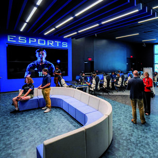 A modern esports facility featuring a large screen, players engaged at computers, and people networking in a comfortable, stylish space.