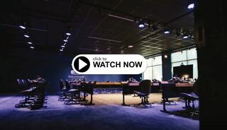 Button with "Click to Watch Now" over a virtual reality learning lab featuring two rows of long tables and chairs at each virtual reality workstation, overhead theater lights and Dreamscape Learn logo on the back wall