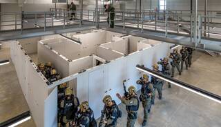 A team of tactical officers in gear navigates a training facility with modular rooms, while another officer observes from a raised platform.