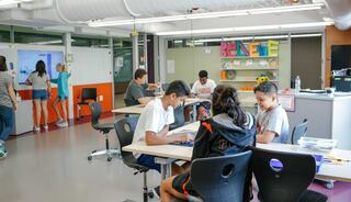 Students are engaged in various activities in a modern classroom, collaborating at desks and writing on interactive boards.