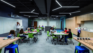 A modern classroom features flexible seating with students engaged in various activities, bright natural light, and interactive learning spaces.