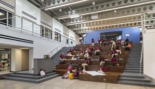 Students sit on large wooden steps in a modern learning space, engaged in various activities, with bright walls and ample natural light.