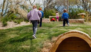 Children play and explore a grassy hillside, climbing and moving among natural surroundings, enjoying a sunny day outdoors.