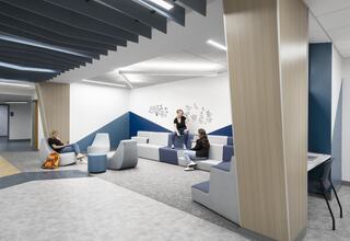 A modern lounge area features stylish seating in gray and blue tones, with people relaxing and studying in a bright, open space.