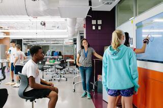 A middle school STEAM classroom filled with students engaged in activities; one girl interacts with a digital screen while a teacher observes.