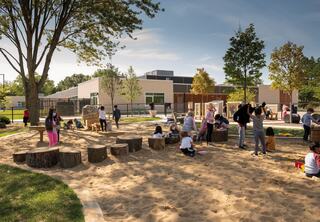 Children play in a sandy area surrounded by trees, wooden logs, and a school building in the background, enjoying a sunny day outdoors.