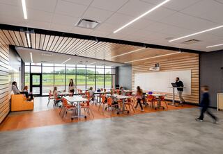 A modern classroom features students seated at tables with orange chairs, a teacher at a podium, and large windows offering outdoor views.