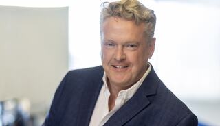 A smiling man with wavy blonde hair wears a dark blazer over a white shirt, posed confidently in a bright, professional setting.