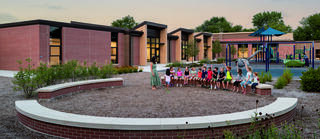 A group of children sits on a circular bench, while a teacher stands nearby. In the background, a playground and modern school buildings are visible.