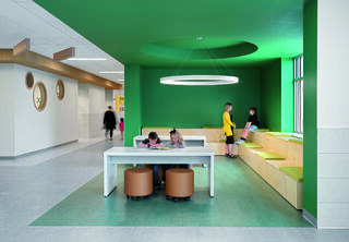 A modern room features vibrant green walls, a large table, light fixtures, and seating areas where children engage in activities.