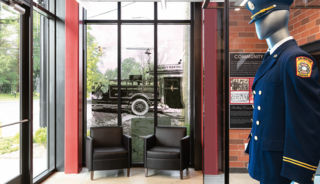 A modern space featuring two black chairs, a mannequin in a blue uniform, and a large window displaying a historical fire truck scene.