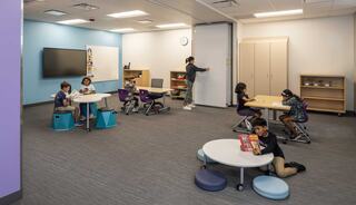 A bright classroom with various seating arrangements, where students engage in activities at tables while others interact nearby.
