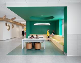 A vibrant room features a large table and cozy seating, where people engage in activities, complemented by a bright green wall and unique lighting.