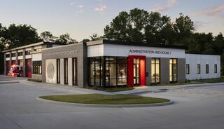 A modern building featuring a sleek design with large windows, labeled "Administration and House 1," surrounded by greenery at sunset.