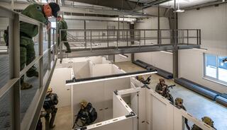 A tactical training facility features officers in gear navigating between white partitions, while others observe from a raised platform.