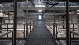 A spacious industrial corridor elevated above a bright, open area, flanked by railings and leading toward a distant light-filled exit.