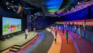 Children gather in a colorful, modern space, following a leader on stage, with vibrant visuals enhancing the lively atmosphere.