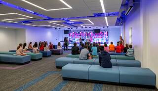 A group of children sits on modern seating, watching a performance in a brightly lit, contemporary space with vibrant lighting and a stage.