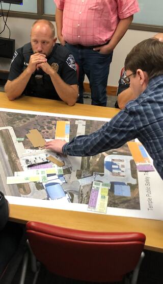 A group discusses a large map during a planning session, with officers and another individual analyzing key areas and details.